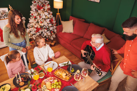 High angle view of happy multi-generation family gathered around the table having Christmas dinner all together at home, grandfather playing the guitar while everybody singing Christmas songsの写真素材