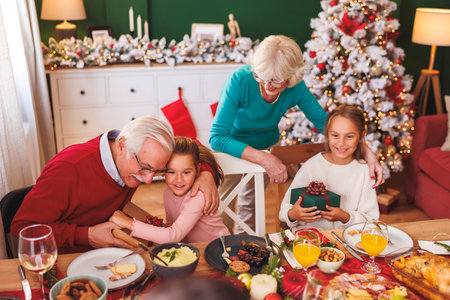 Happy multi-generation family gathered around the table having Christmas dinner all together at home, grandparents surprising granddaughters with Christmas presentsの写真素材