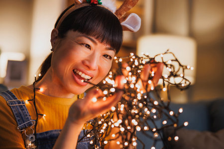 Beautiful Japanese woman wearing costume reindeer antlers arranging Christmas lights while decorating home for Christmas eve celebrationの写真素材