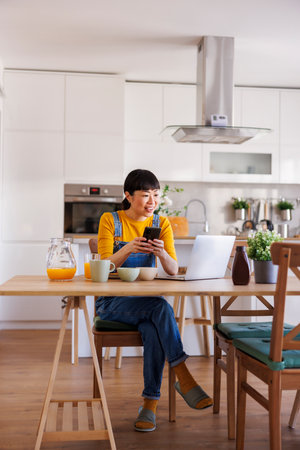 Mature Asian woman typing text message using smart phone and having breakfast while working remotely from home using laptop computerの写真素材