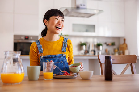Beautiful mature Asian woman having breakfast at home adding fresh berries to wafflesの写真素材