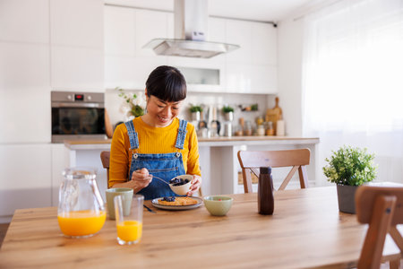 Beautiful mature Asian woman having breakfast at home adding fresh berries to wafflesの写真素材