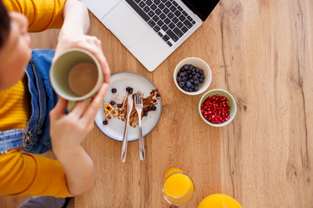High angle view of female freelancer working remotely from home using laptop computer drinking coffee and finishing up her breakfastの写真素材