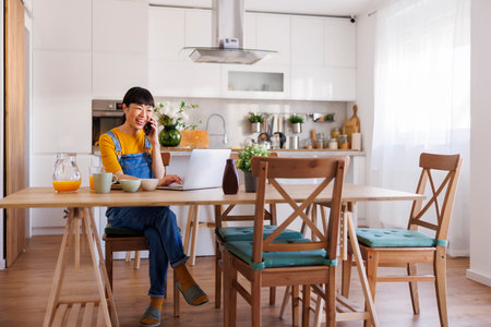 Mature Asian woman having phone conversation using smart phone and having breakfast while working remotely from home using laptop computerの写真素材