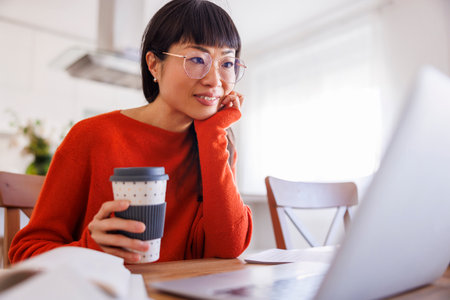 Mature Asian woman drinking coffee while working remotely from home using laptop computerの写真素材
