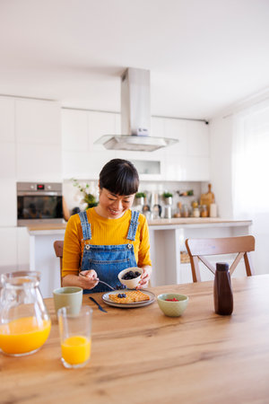 Beautiful mature Asian woman having breakfast at home adding fresh berries to wafflesの写真素材