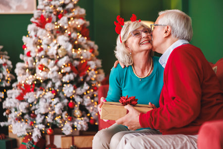 Senior couple relaxing and enjoying spending Christmas day together at home exchanging presents by the Christmas treeの写真素材