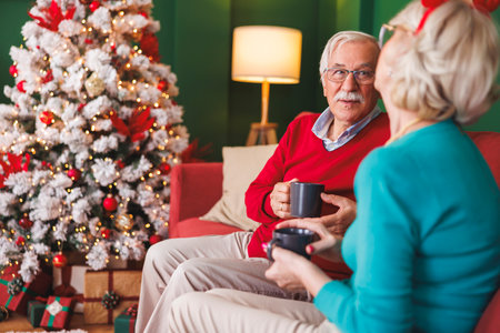 Happy senior couple enjoying spending Christmas day together at home, drinking coffee and having a conversation by the Christmas treeの写真素材