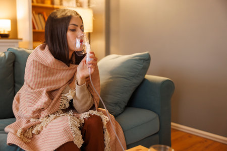 Sick woman having flu and fever sitting on sofa wrapped in blanket using inhaler to treat respiratory infectious diseaseの写真素材