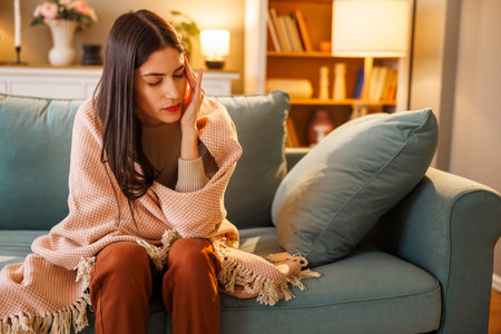 Sick young woman having flu and fever sitting on living room sofa wrapped in blanket, holding head in hands having headacheの写真素材