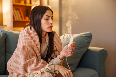 Sick woman having flu and fever sitting on sofa wrapped in blanket using inhaler to treat respiratory infectious diseaseの写真素材