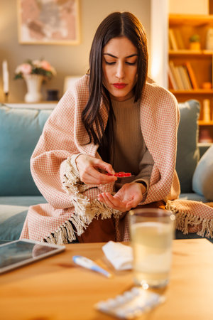 Sick woman having flu and fever sitting on sofa at home wrapped in blanket, taking pillsの写真素材