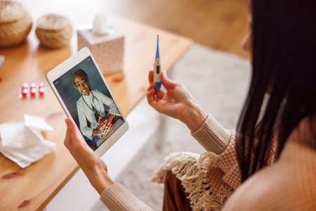 Sick woman having flu and fever sitting on sofa at home wrapped in blanket, having online doctor's appointment using tablet computerの写真素材