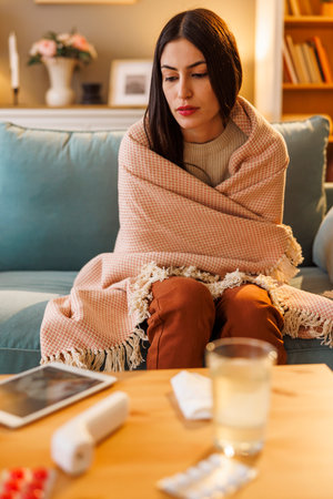 Sick woman having flu and fever sitting on sofa wrapped in blanket while having online doctor's checkup using tablet computerの写真素材