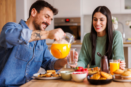 Beautiful couple relaxing and having breakfast pouring fresh orange juice and enjoying their leisure time at homeの写真素材