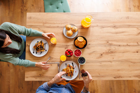 Top view of beautiful couple in love adding chocolate topping on waffles while having breakfast at home, relaxing and enjoying their leisure time togetherの写真素材