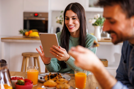 Beautiful couple in love having breakfast at home, using tablet computer, relaxing and enjoying their time togetherの写真素材