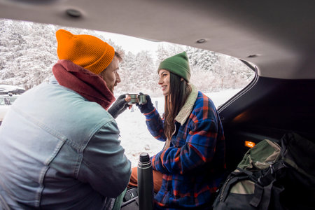 Beautiful couple in love sitting in a car trunk taking a break while going for a winter vacation road trip, drinking hot tea and enjoying snowy day in natureの写真素材