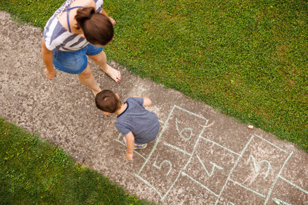 High angle view of mother and cute toddler son having fun playing hopscotch game, hopping on one leg...