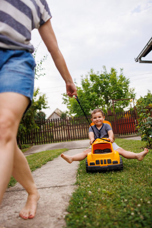 Mother having fun pushing toddler son sitting in trailer of large plastic toy truck and spraying water with gun, playing together in the backyard of their homeの写真素材