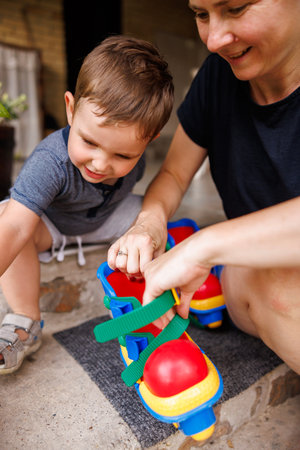 Mother and toddler son having fun playing in the backyard of their home, mother helping child to put...