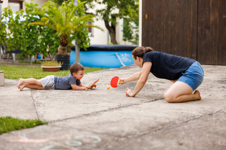 Mother and toddler son having fun playing with table tennis rackets and ball while spending time together in the backyard of their homeの写真素材