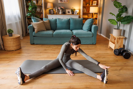 High angle view of active fit young woman in sportswear sitting on exercise mat stretching out while...