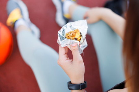 High angle view of young woman in sportswear sitting on an outdoor basketball court eating an energy bar while taking workout breakの写真素材