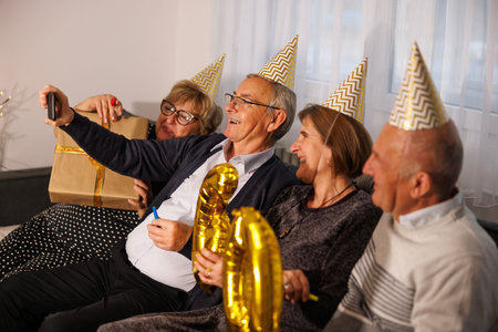 Group of cheerful senior friends gathered at friend's 60th birthday party, having fun wearing party...