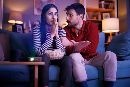 Couple having fun watching football game on TV and cheering for their team while relaxing at home at...