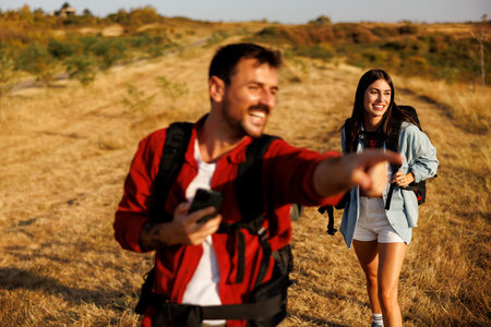 Active couple carrying backpacks having fun hiking in nature on sunny summer day, man using smart...