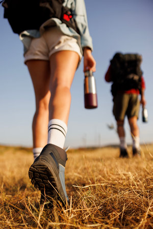 Back view of detail of couple carrying backpacks and water bottles hiking in nature, walking away...
