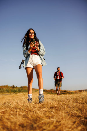 Active young couple carrying backpacks having fun hiking in nature on sunny summer day, woman using...