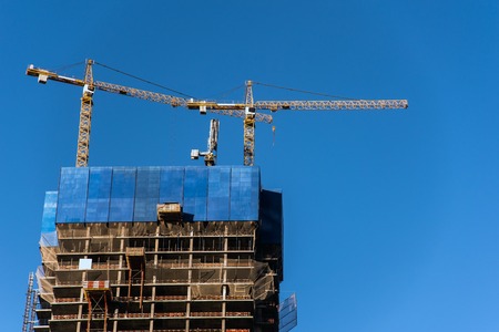 Industrial construction tower cranes and high-rise building under construction against blue skyの写真素材