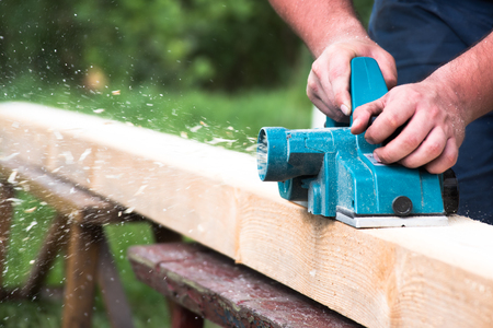 Close up hands of carpenter working with electric planer on wooden plankの写真素材