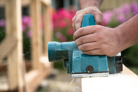 Close up hands of carpenter working with electric planer on wooden plank in carpentry workshopの写真素材
