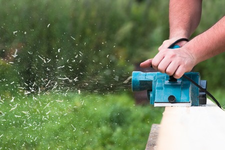 Close up hands of carpenter working with electric planer on wooden plank in carpentry workshopの写真素材