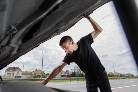 Closeup portrait, young man having trouble with his broken auto, opening hood trying to fix engine, isolated green trees outside background.の写真素材