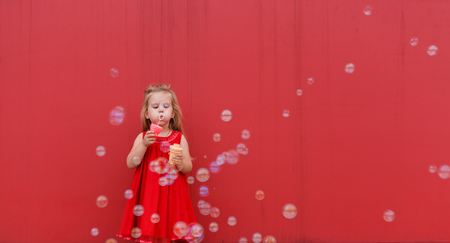 little girl in red drtess blowing soap bubbles on the red backgroundの写真素材