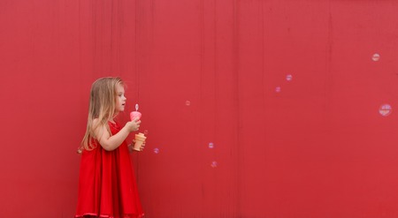 little girl in red drtess blowing soap bubbles on the red backgroundの写真素材