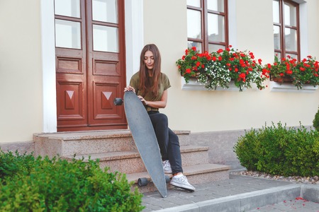 Stylishly dressed woman sitting on the steps with longboard in the hands of front of the house.の写真素材