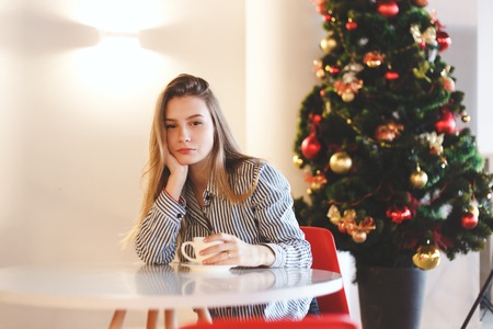 Young beautiful woman drinks hot coffee in cafe with Christmas tree decorationsの写真素材