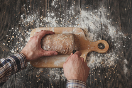 Baking and cooking concept background. Hands of baker closeup cutting bread loaf with knife on rustic wooden table sprinkled with flour.の写真素材