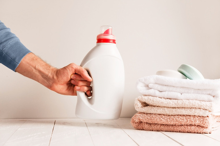 Mans hand putting on the table a jar of gel for washing clothes. Against the background of a pile of towels and soapの写真素材