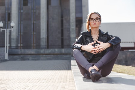 Street fashion concept - stylish cool girl in rock black style sitting among urban background outdoorの写真素材