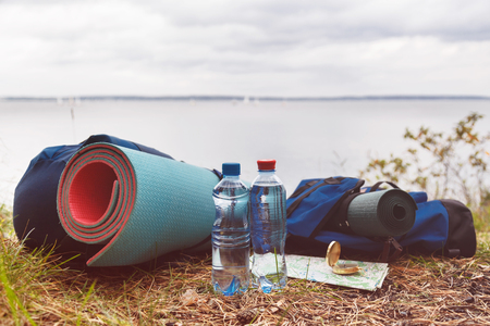 A set of tourist accessories necessary for tourism in the wild. Bottled water, rugs, backpacks, a map and a compass on the grass in the forest on the shoreの写真素材