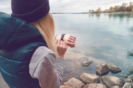 Sportswoman uses an electronic bracelet-pedometer standing on the lake shore. Travel and healthy lifestyle concept. Back viewの写真素材