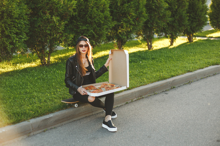 Young hipster woman with skateboard has a lunch with slice of pizza sitting outdoorの写真素材