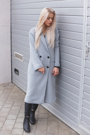Hipster girl wearing gray coat posing against rough street wall, minimalist urban clothing styleの写真素材