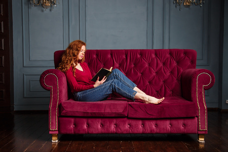 Rich young redhead woman reading a book on red velvet sofaの写真素材